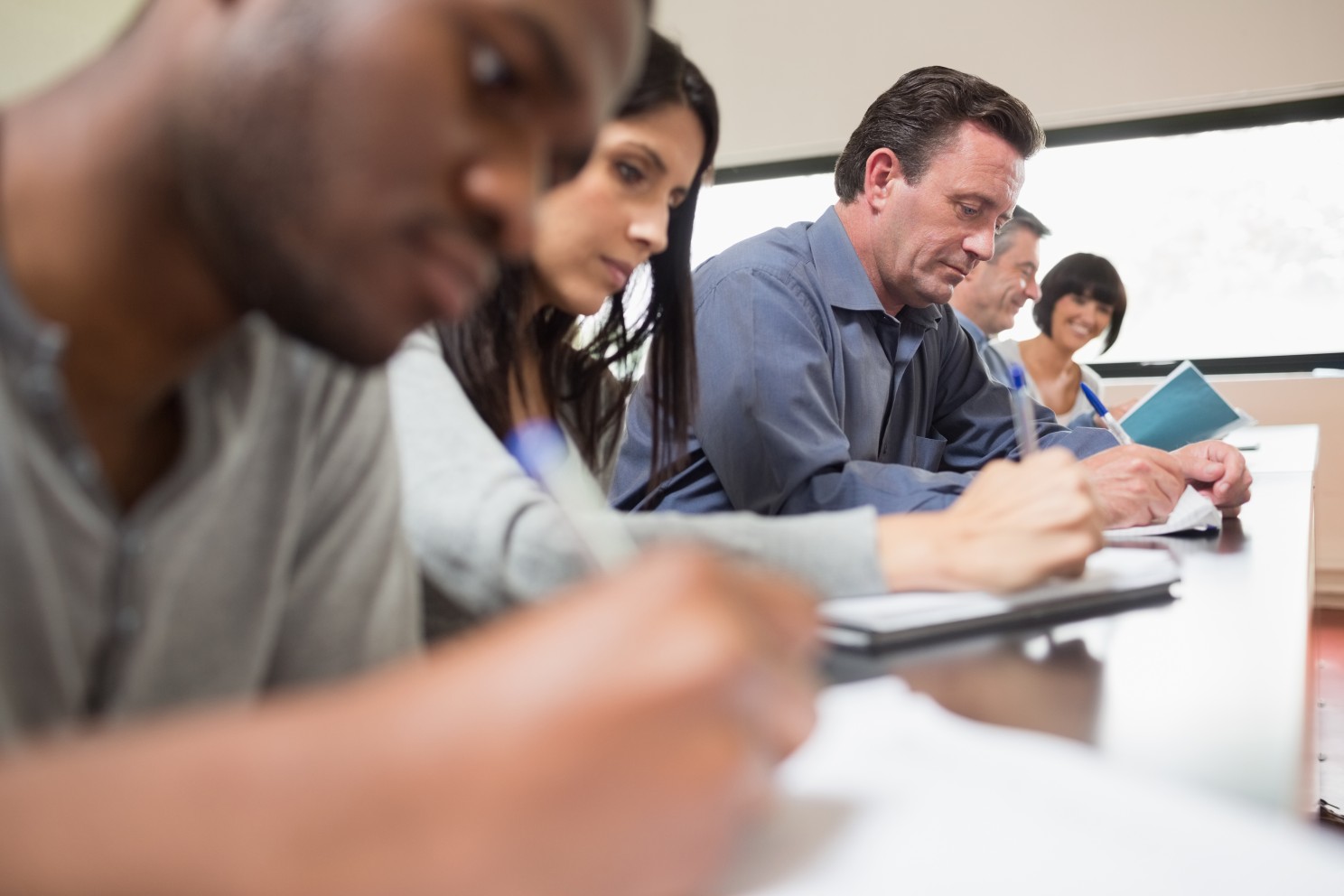 An image of five people sitting in a row in front of a table writing in notepads using blue pens.