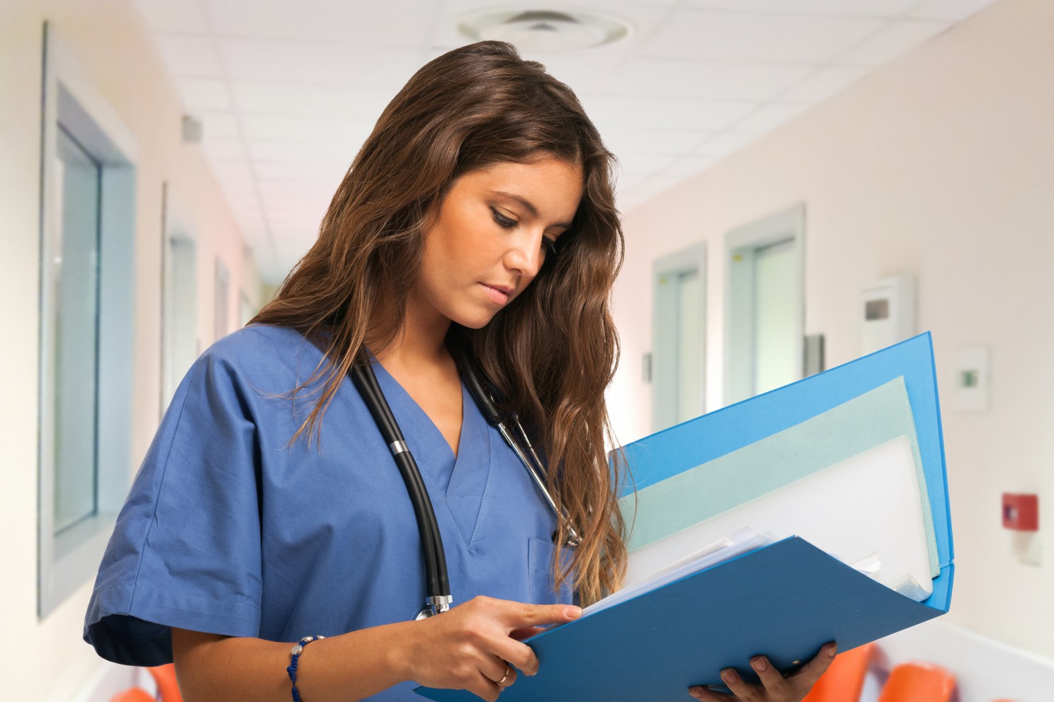 A nurse with long brown wavy hair wearing blue scrubs standing in a hallway reading a patient file.